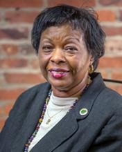 The image shows a woman (Manervia) posed for a professional portrait. She’s wearing a dark blazer over a light top, along with a necklace and a small circular pin on her lapel. Her hair is short and neatly styled, and she’s smiling slightly. The background appears to be a brick wall, giving the photo a warm, formal-yet-approachable feel.